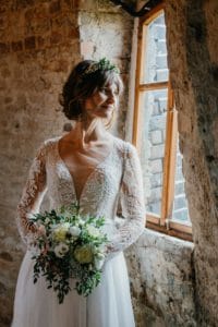 Braut schaut aus Fenster Spitzenkleid florale Elemente Blumenkranz